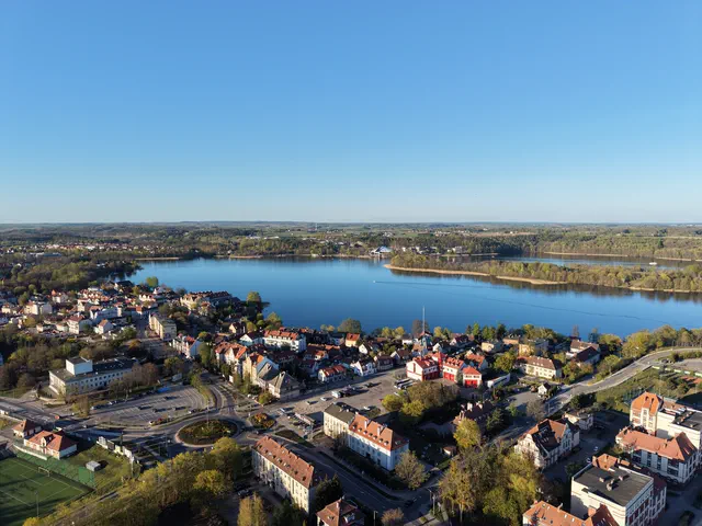 Ujęcie z drona na dachy domów nad jeziorem i zabudowę na przeciwległym brzegu.